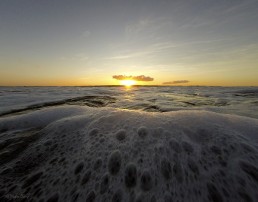 Waves whip up bubbles as the sun sets on a distant horizon.