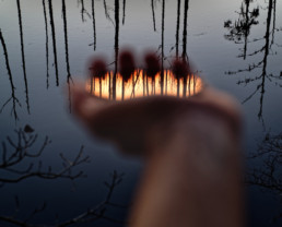 A blurry hand outstretched in front of darkening waters hold a filter (almost invisible) with a clearly visible reflection of tree trunks and sunset colors.